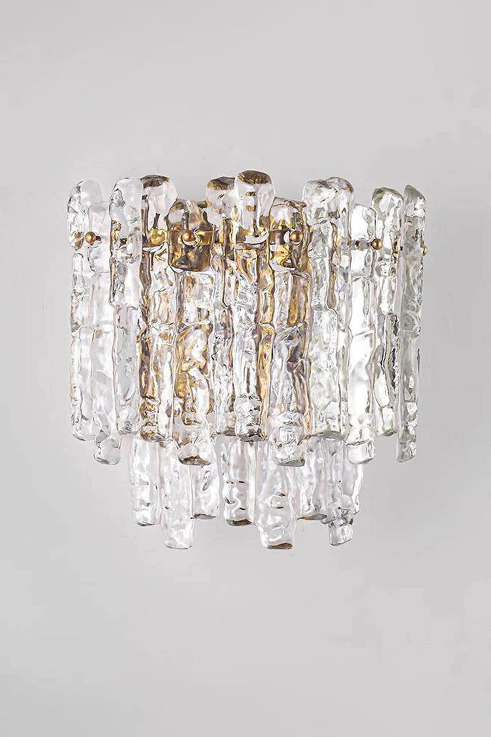 Elara Textured Cast Glass Wall Lamp | Textured Cast Glass, Polished Brass | Medium angle shot of the illuminated lamp against a neutral grey wall, demonstrating its shape and the dramatic textured glass effect in a corridor or entryway | vexa lamps