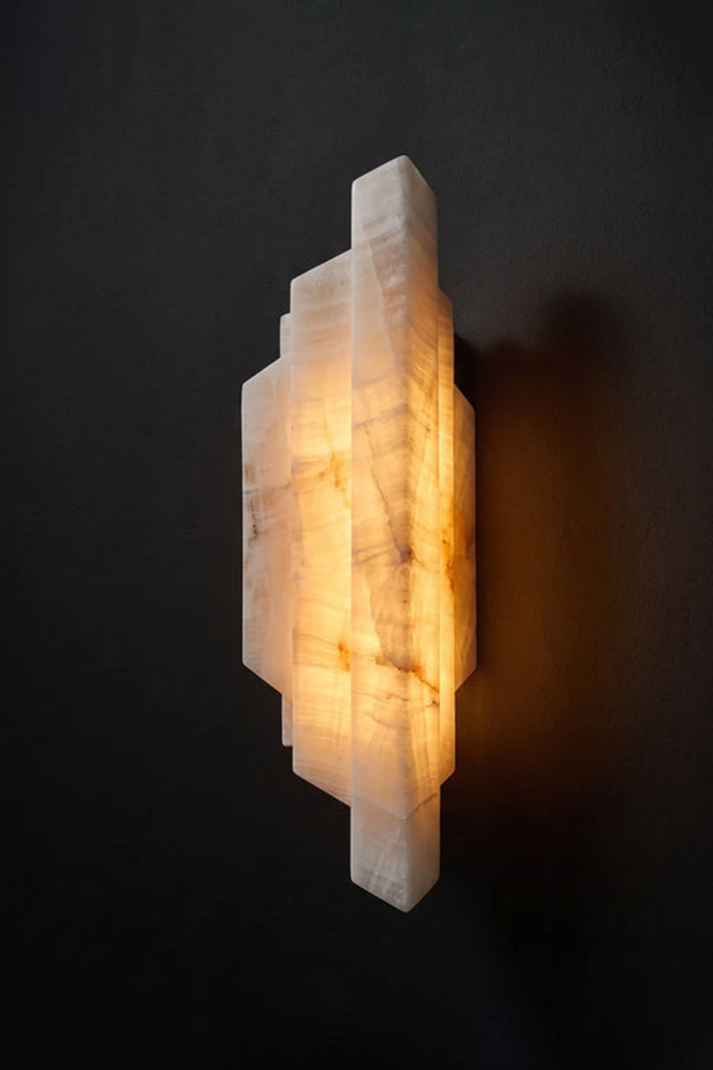 Alaric Tiered Alabaster Wall Sconce | Natural Alabaster and Metal | Angled side view of the illuminated multi-tiered geometric stone fixture against a dark black background | vexa lamps