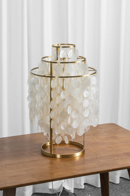 Aurelia Tiered Capiz Table Lamp | Capiz Shell, Metal | Staged unlit view of the tiered shell table lamp sitting on a rich wood surface next to sheer white blinds | vexa lamps