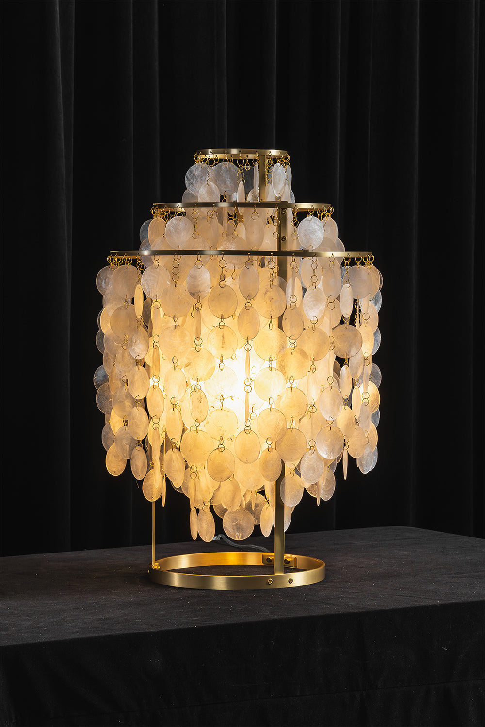 Aurelia Tiered Capiz Table Lamp | Capiz Shell, Metal | Dramatic dark studio shot of the fully illuminated tiered lamp radiating an intense golden glow against a black backdrop | vexa lamps