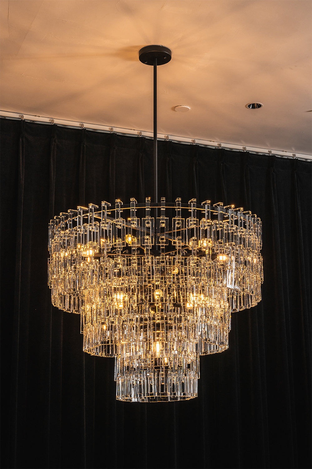 Aven Tiered Glass Prism Chandelier | Glass, Metal | Full view of the illuminated tiered crystal fixture casting a soft golden glow against a dramatic dark wall backdrop | vexa lamps