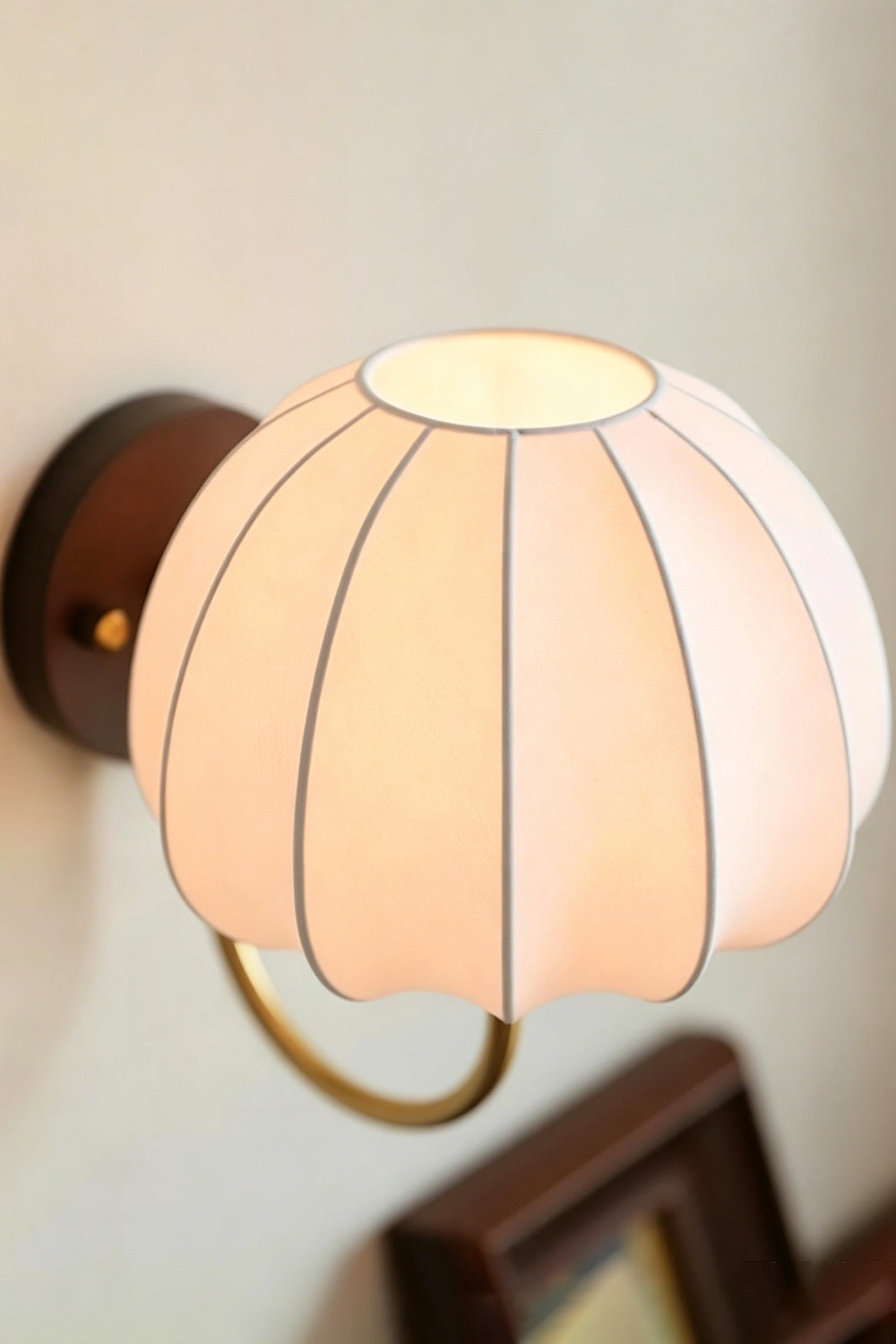Baird Mid Century Pumpkin Wall Lamp | Metal, Fabric, Wood | High-angle shot of the fully lit pumpkin sconce mounted over a wooden picture frame | vexa lamps