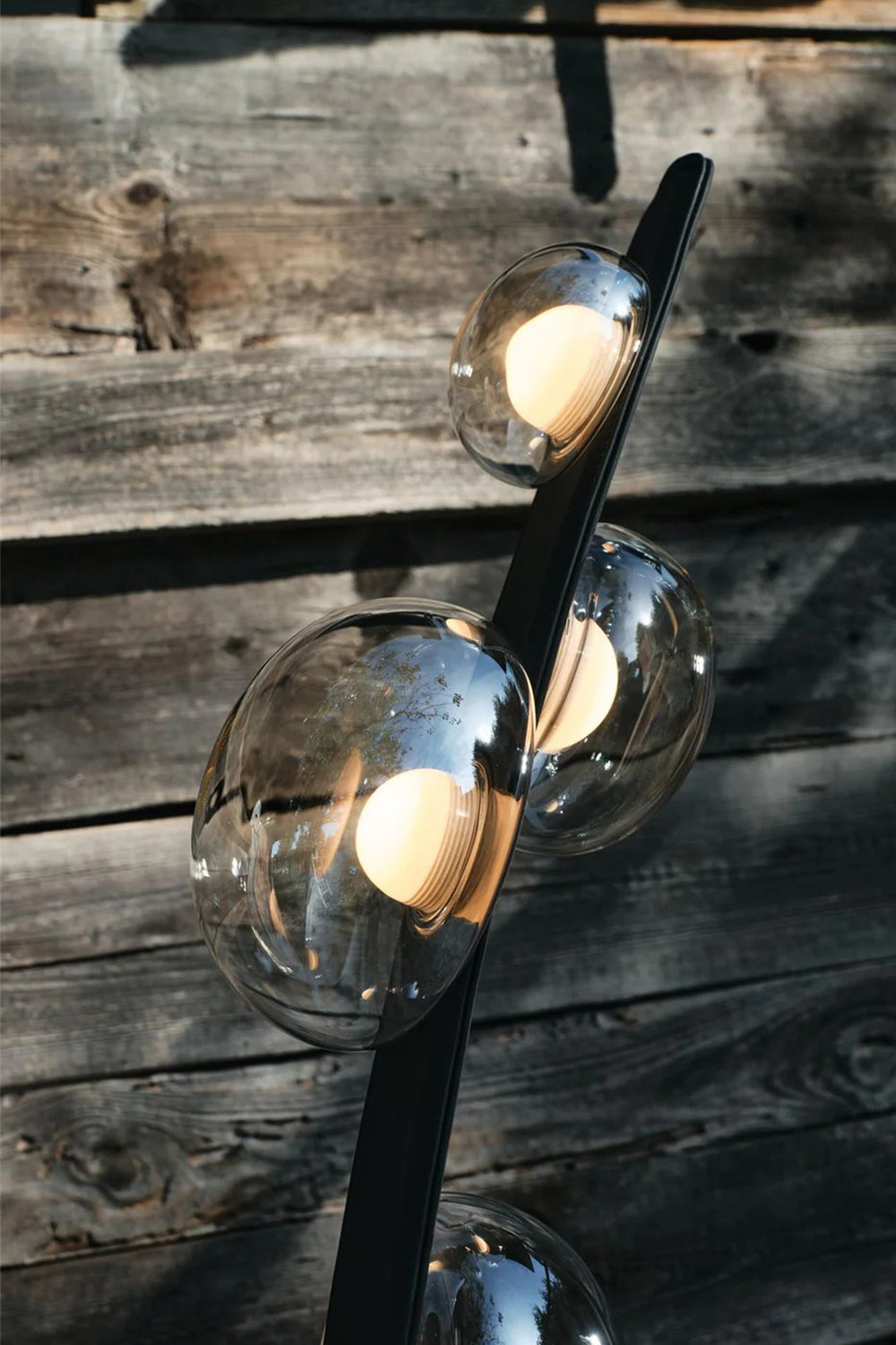 Drosia Dewdrop Floor Lamp | Glass, Metal | Close-up focus on the fully illuminated warm globe bulbs refracting light through the clear dewdrop glass shades against a rustic dark wooden wall | Detail shot | vexa lamps