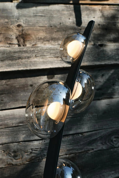 Drosia Dewdrop Floor Lamp | Glass, Metal | Close-up focus on the fully illuminated warm globe bulbs refracting light through the clear dewdrop glass shades against a rustic dark wooden wall | Detail shot | vexa lamps