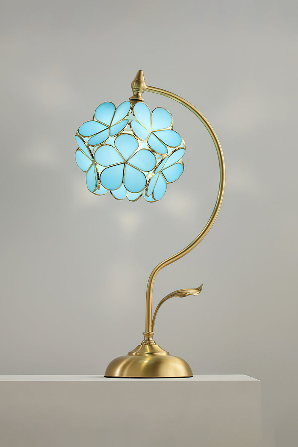 Evanthe Blossom Stained Glass Table Lamp | Stained Glass, Metal | Studio front view of the vibrant sky blue variant fully illuminated on a white pedestal block against a grey wall | vexa lamps
