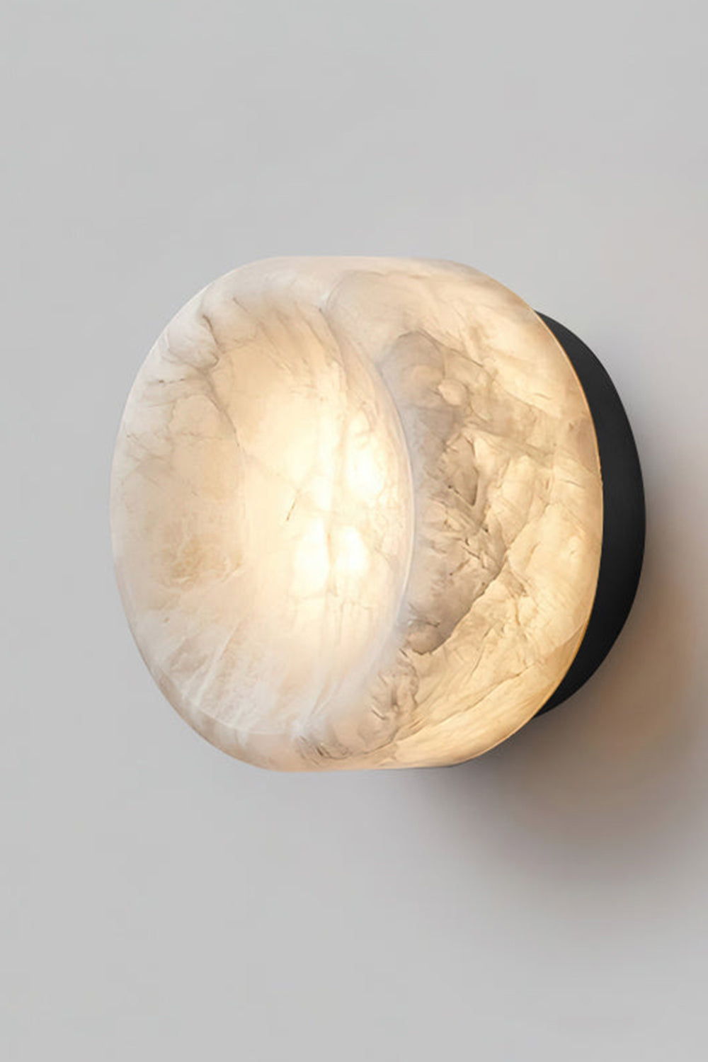 Isolde Concave Alabaster Disc Wall Sconce | Natural Alabaster and Black Metal | Studio close-up shot of the lit sconce featuring a matte black finish base on a light wall | vexa lamps