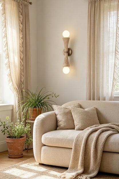 Katia Travertine Hourglass Wall Lamp | Travertine, Metal | Positioned gracefully on a living room wall, radiating soft accent lighting next to a plush beige fabric sofa with throw blankets | vexa lamps