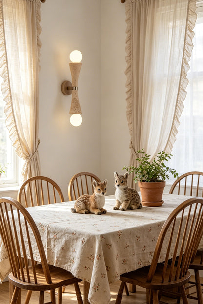 Katia Travertine Hourglass Wall Lamp | Travertine, Metal | Illuminating a rustic cottage dining area, mounted on the wall near a dining table set with a cloth and wooden spindle back chairs | vexa lamps