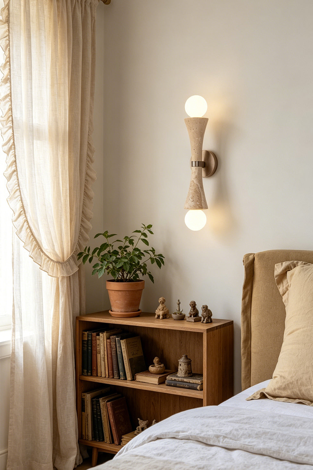 Katia Travertine Hourglass Wall Lamp | Travertine, Metal | Providing a warm ambient light in a cozy bedroom corner next to a wooden storage shelving unit and linen drapery window | vexa lamps