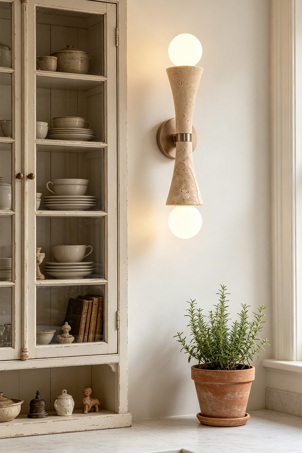 Katia Travertine Hourglass Wall Lamp | Travertine, Metal | Styled in a contemporary interior next to a vintage glass hutch cabinet filled with ceramic dishes and a terracotta plant pot | vexa lamps
