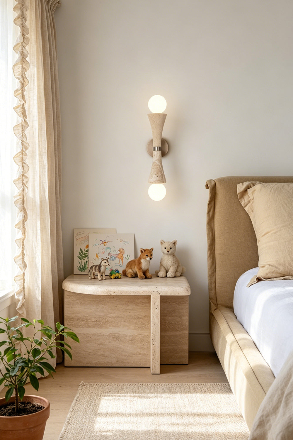 Katia Travertine Hourglass Wall Lamp | Travertine, Metal | Mounted as an elegant bedside reading sconce casting an upward and downward warm glow next to a wooden nightstand and a cozy platform bed | vexa lamps