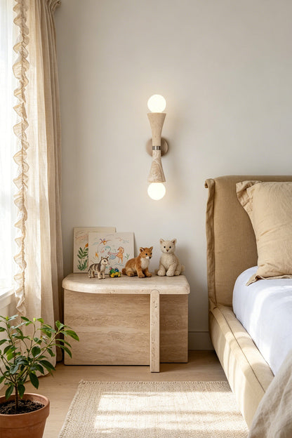 Katia Travertine Hourglass Wall Lamp | Travertine, Metal | Mounted as an elegant bedside reading sconce casting an upward and downward warm glow next to a wooden nightstand and a cozy platform bed | vexa lamps