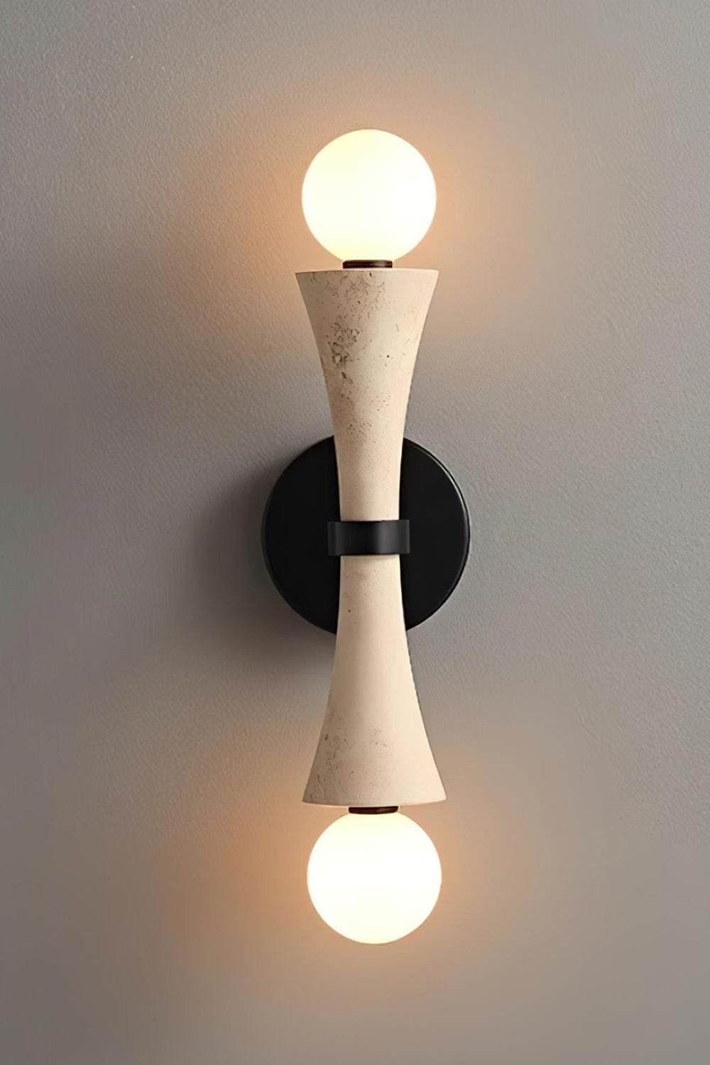 Katia Travertine Hourglass Wall Lamp | Travertine, Metal | Studio front view of the fully illuminated hourglass stone wall fixture casting a brilliant dual-directional warm light halo against a grey backdrop | vexa lamps