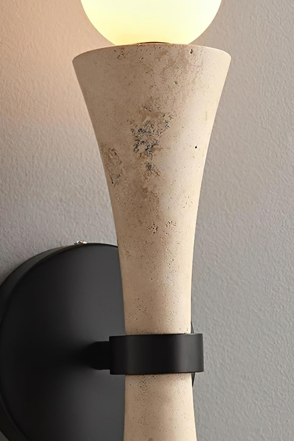Katia Travertine Hourglass Wall Lamp | Travertine, Metal | Side profile up-close view showing the textured beige stone hourglass body held firmly by a matte black metallic ring clamp collar | Detail shot | vexa lamps