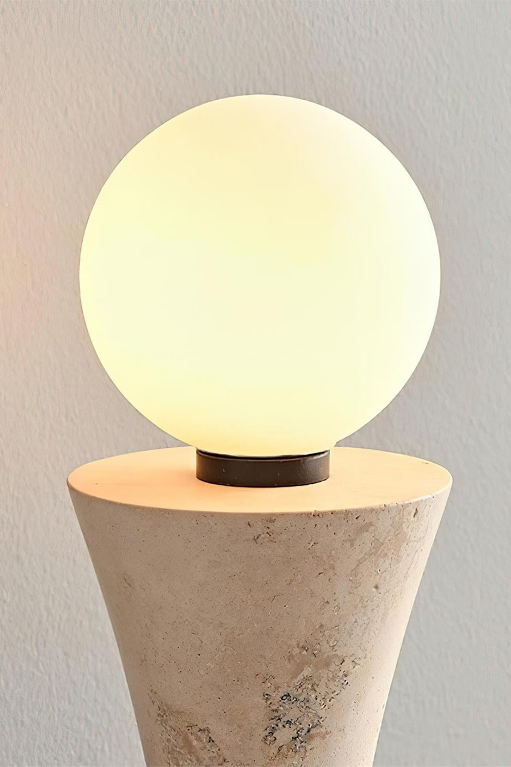 Katia Travertine Hourglass Wall Lamp | Travertine, Metal | Detailed shot of the top spherical globe light bulb illuminated, radiating a soft warm white glow over the stone texture | Detail shot | vexa lamps