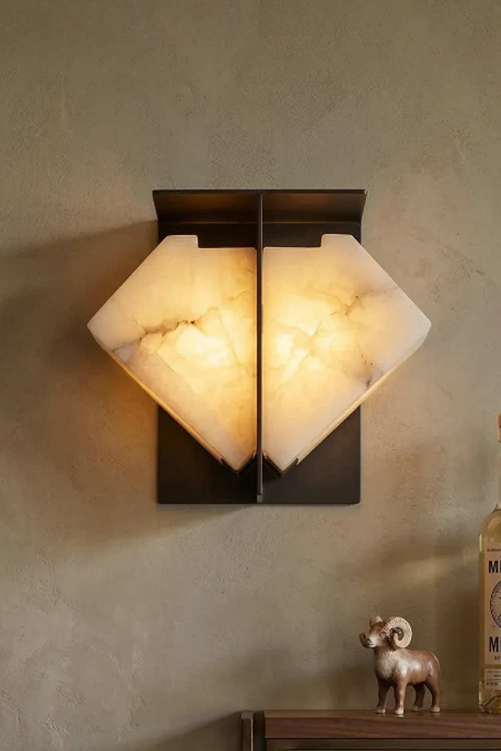 Lysandra Alabaster Folded Halo Wall Lamp | Cozy Mood Lighting | Fully lit geometric stone wall light casting an inviting yellow halo on a textured plaster wall above a shelf decoration | vexa lamps