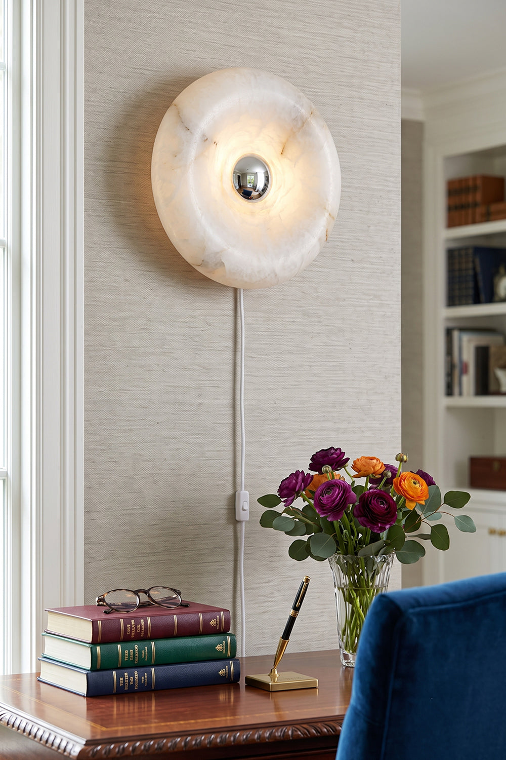 Ondine Ring Alabaster Wall Sconce illuminating a cozy room with books and flowers.