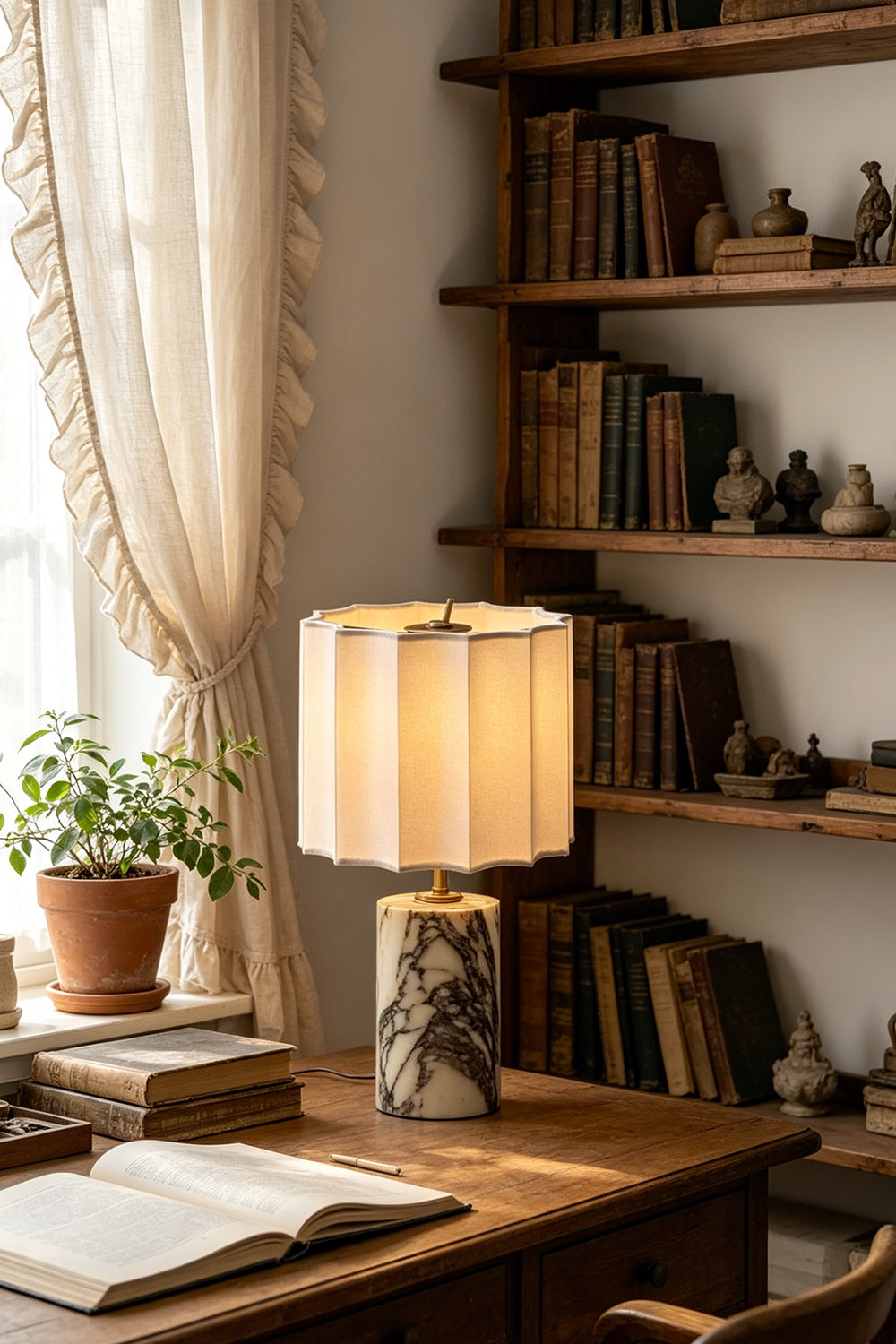Orbis Veil Pleated Marble Table Lamp on a desk with books, surrounded by greenery and warm lighting in a cozy room.