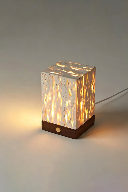Radiant Monolith Travertine Table Lamp | Travertine, Wood | Lit angled view on grey surface | Intricate internal light pattern | Dark wood base, warm glow | vexa lamps