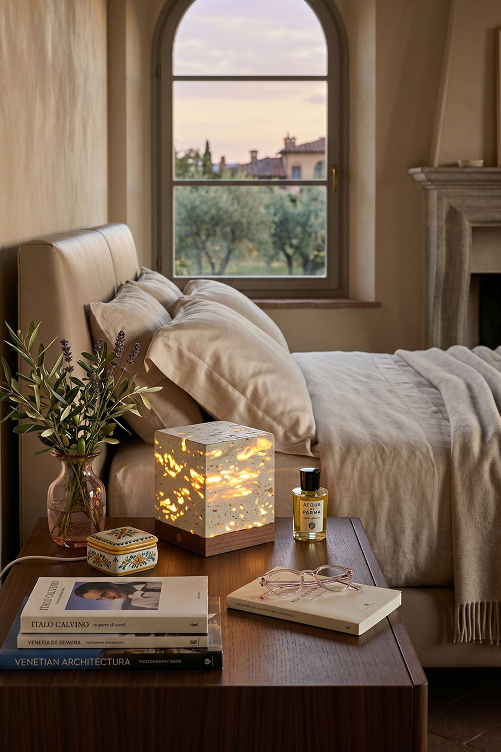 Radiant Monolith Travertine Table Lamp | Travertine, Wood | Lit on wood nightstand | Tuscan bedroom landscape view | Arched window frame, hills | vexa lamps