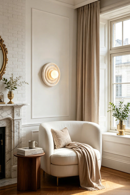 Rhea Rippled Alabaster Wall Sconce | Cozy Reading Nook | Mounted elegantly on a white wall casting a soft warm golden halo over a plush cream armchair and wooden side table in a classical room layout | vexa lamps