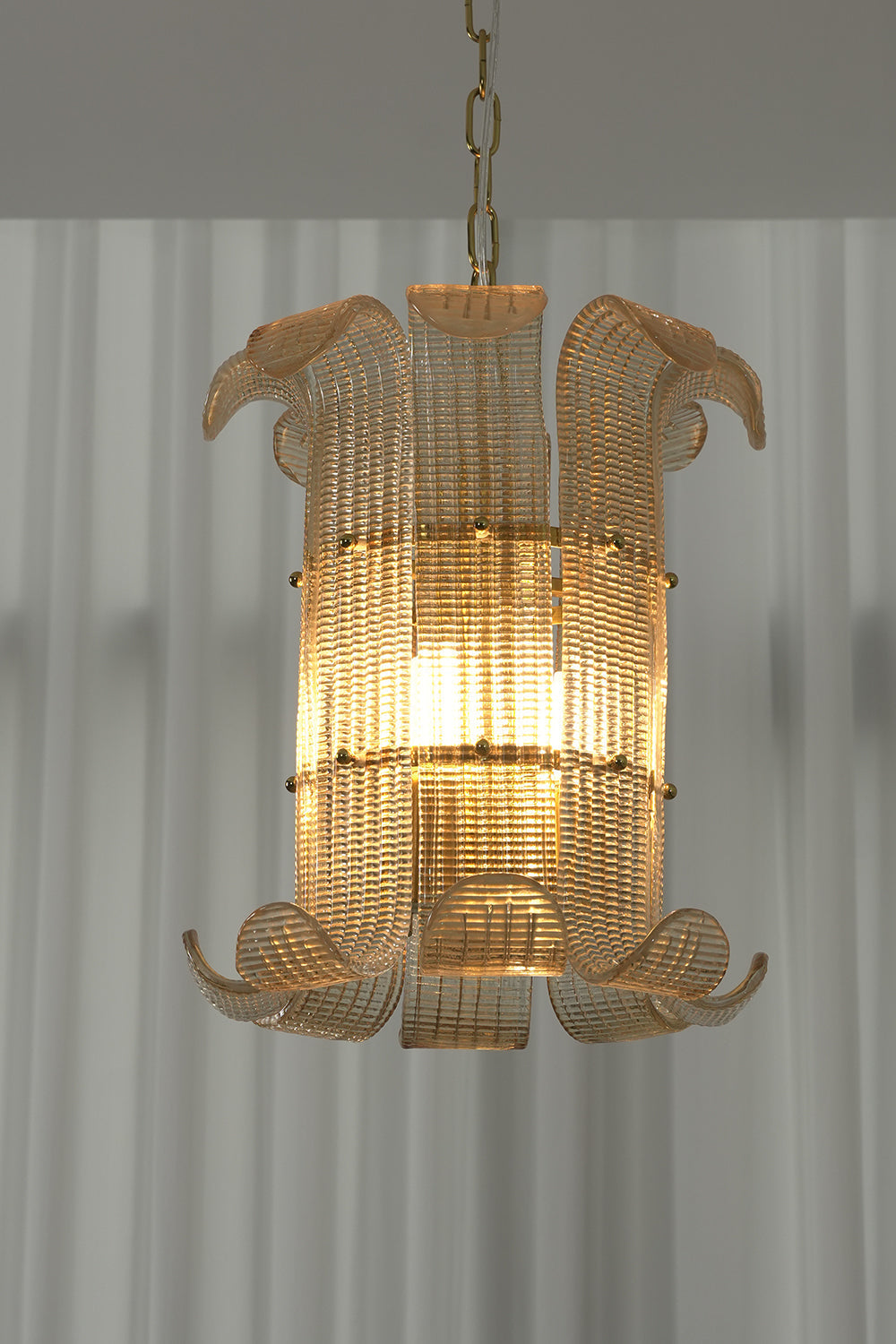 Lit ribbed glass scroll chandelier against white curtain | Metal | Glass | Front view showing the elegant scroll shape and warm glow | vexa lamps