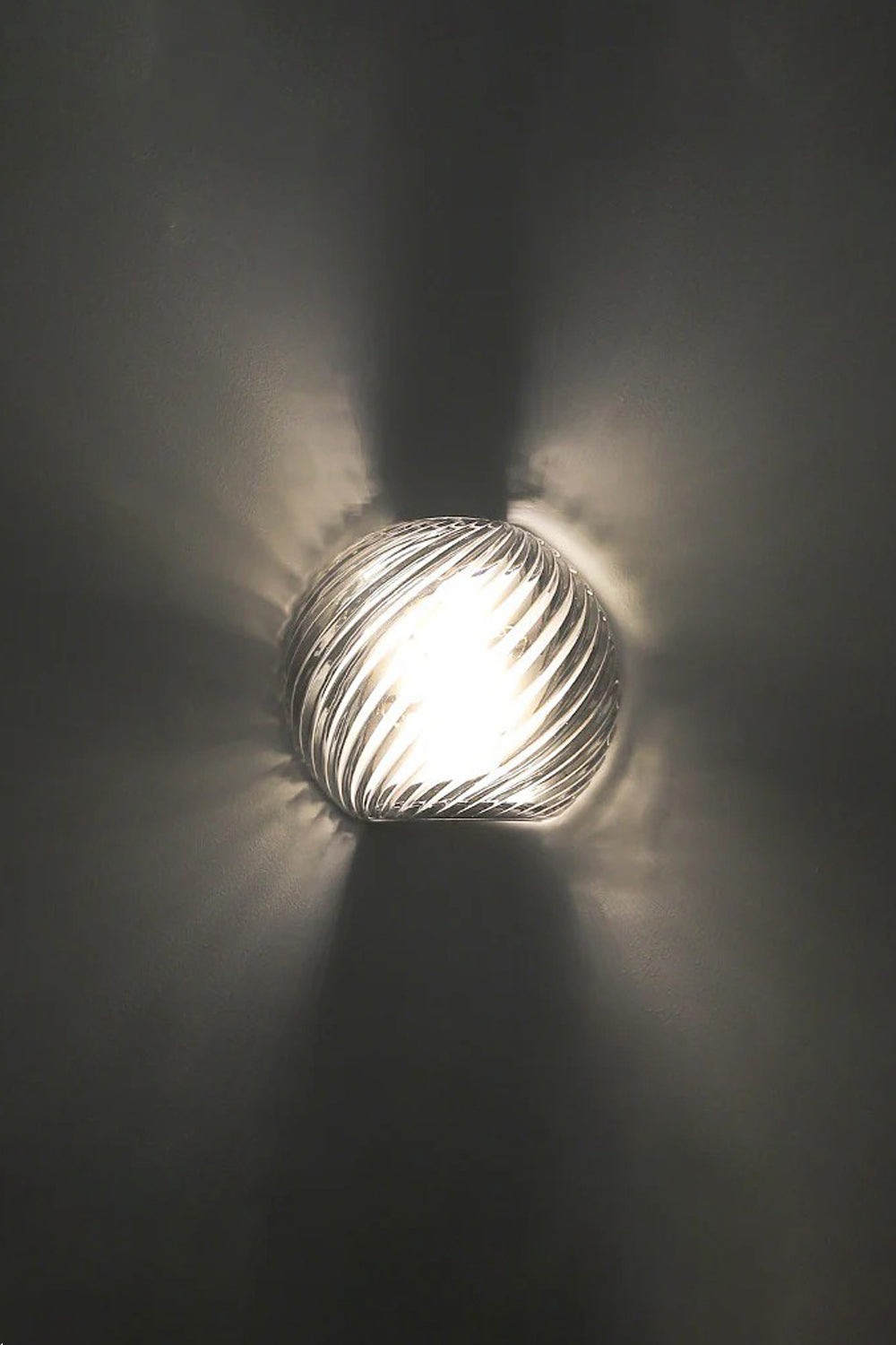 Silas Ribbed Glass Wall Sconce | Ribbed Glass, Metal | Close-up illuminated view against a dark charcoal backdrop, showing the dramatic starburst ribbed light patterns cast on the wall | vexa lamps