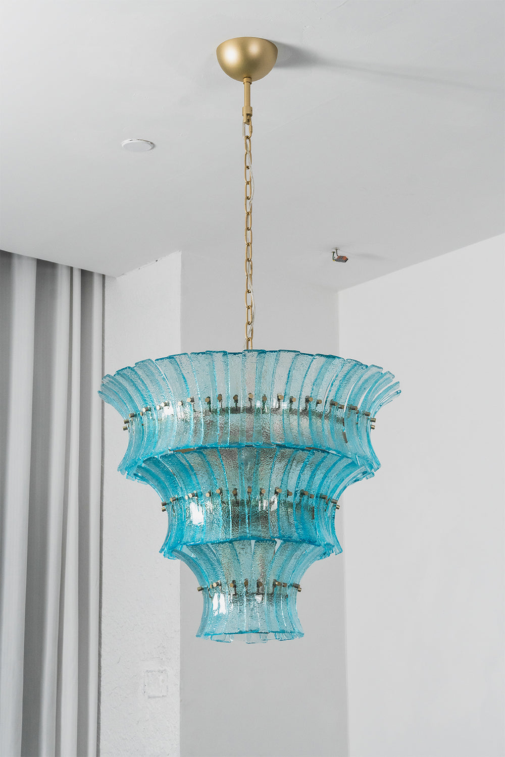 Thalassa Aquamarine Textured Glass Chandelier | Glass, Metal | Studio staged view of the tall multi-tiered lit aquamarine glass chandelier casting a rich sea-colored glow near grey window curtains | vexa lamps
